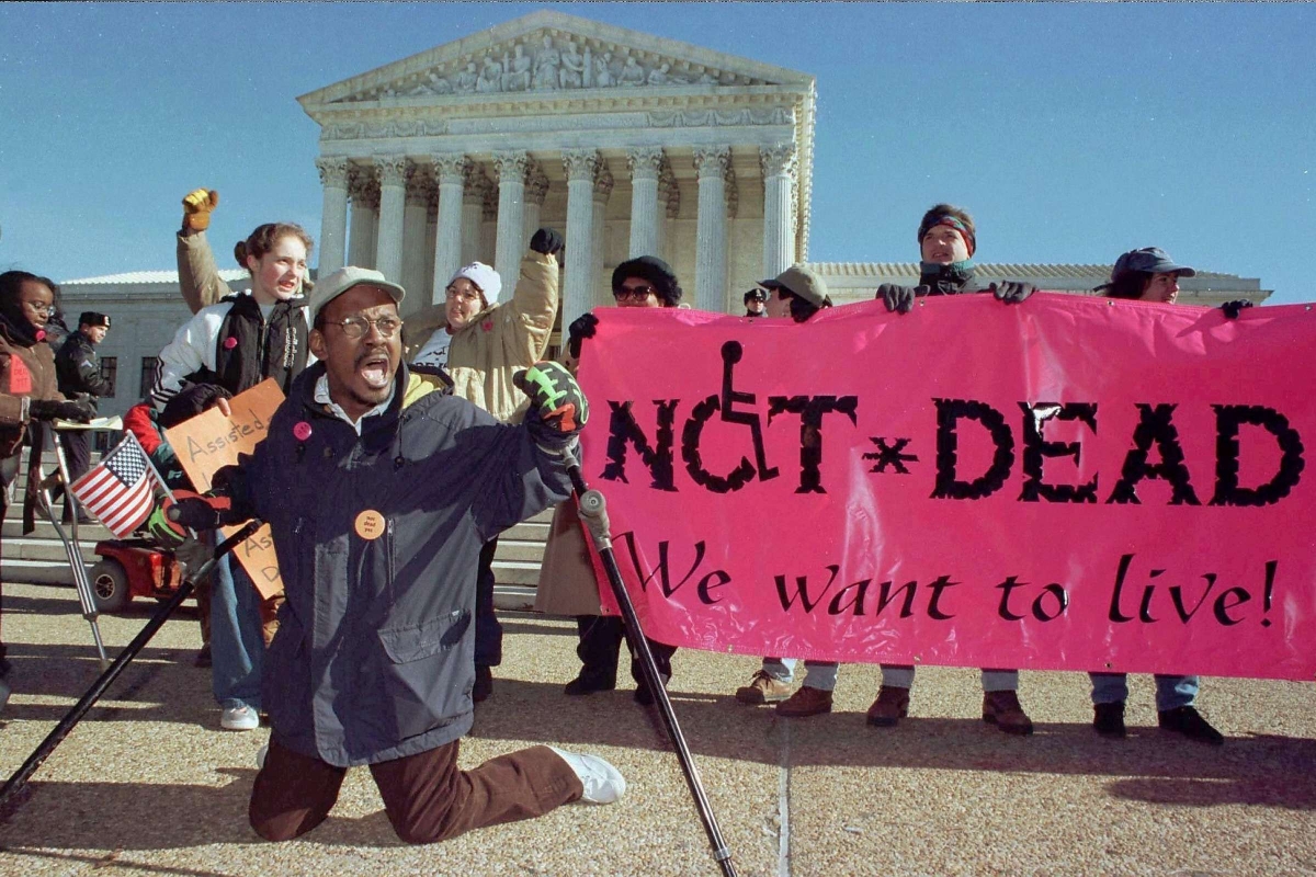 A Black man leads a group of protesters in front of the Supreme Court, kneeling with his crutches outspread and his mouth mid-yell, while holding a small American flag. A neon pink banner to the right reads, "NOT DEAD. We want to live!"