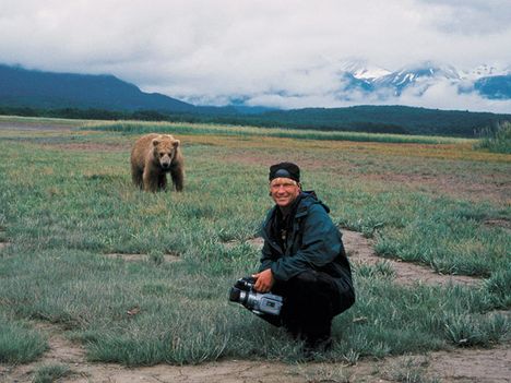 A man squats in front of a field with flowers and mountains in the background, holding a video camera, with a grizzly bear in the bacground