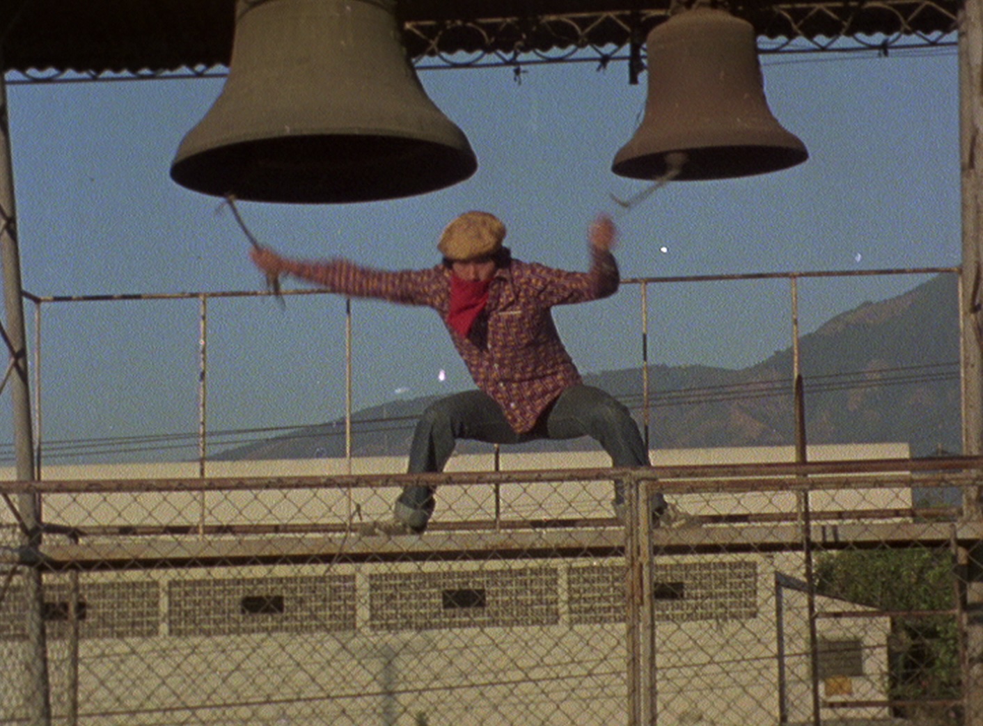 Perched on a thin ledge, a man in a checkered shirt rings two bells.