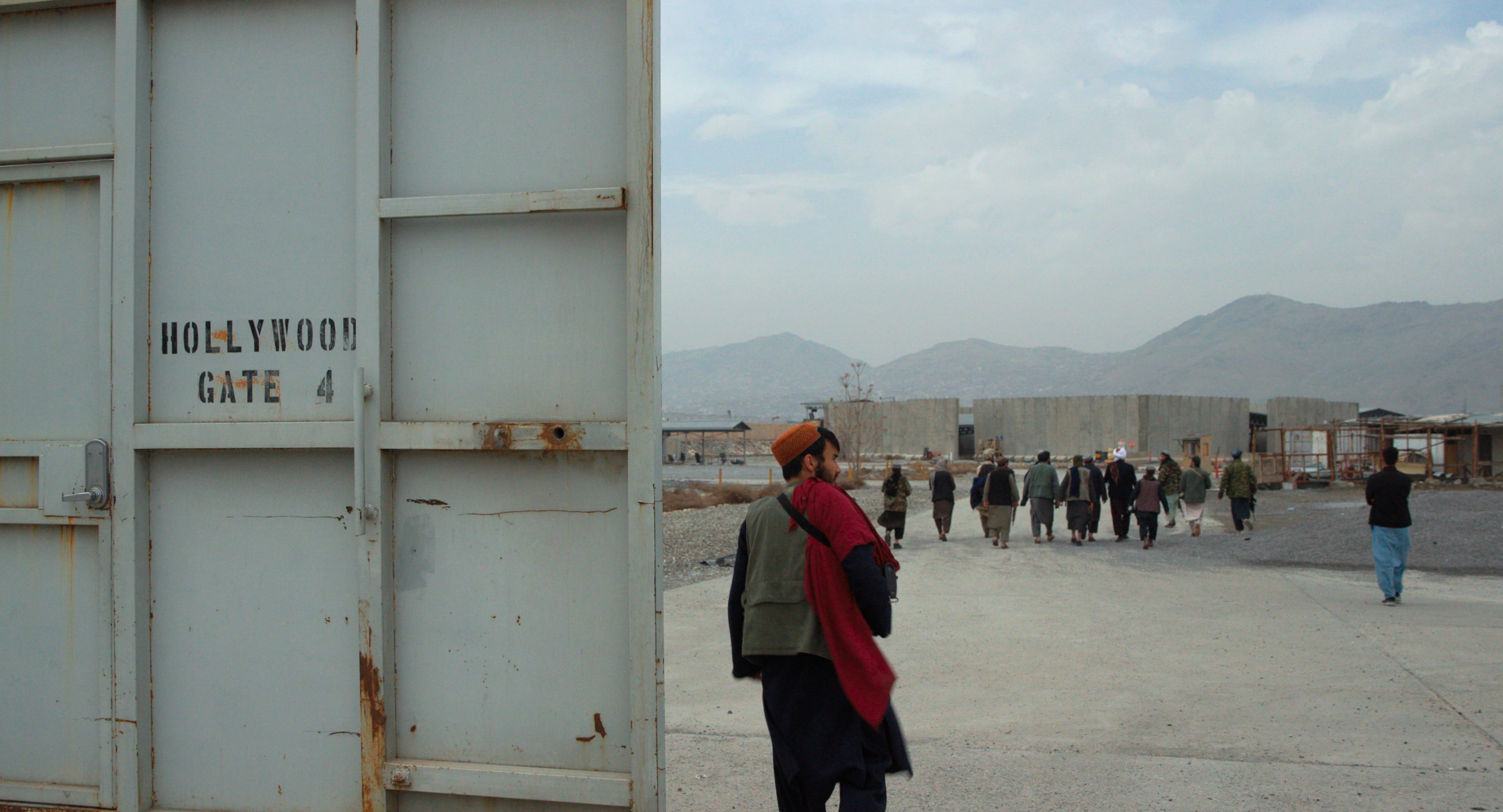 A man stands in the foreground holding a gun. The sign next to him reads ‘Hollywood Gate’. A row of people are in the background, appearing to be monitored by the armed man.