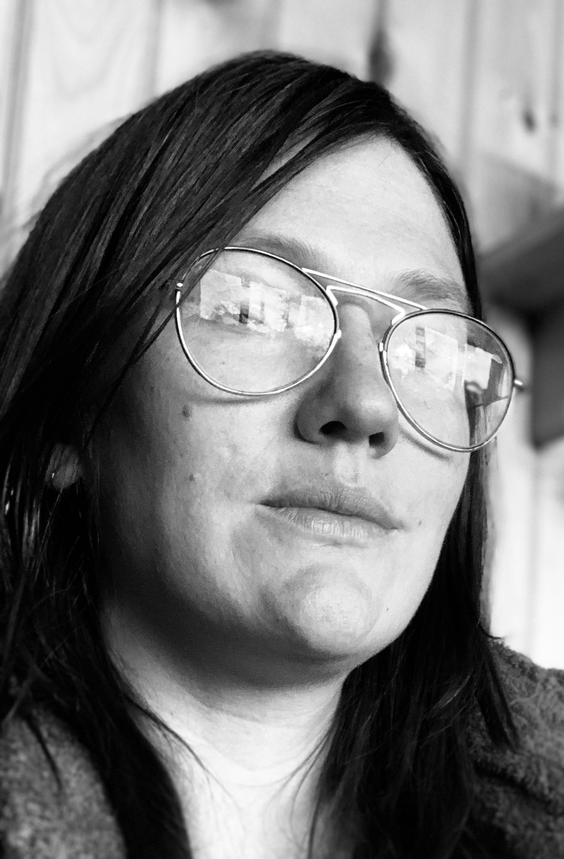 A black-and-white, close-up portrait of a white woman with black hair, wearing thin-framed glasses that have windows in the reflection. 