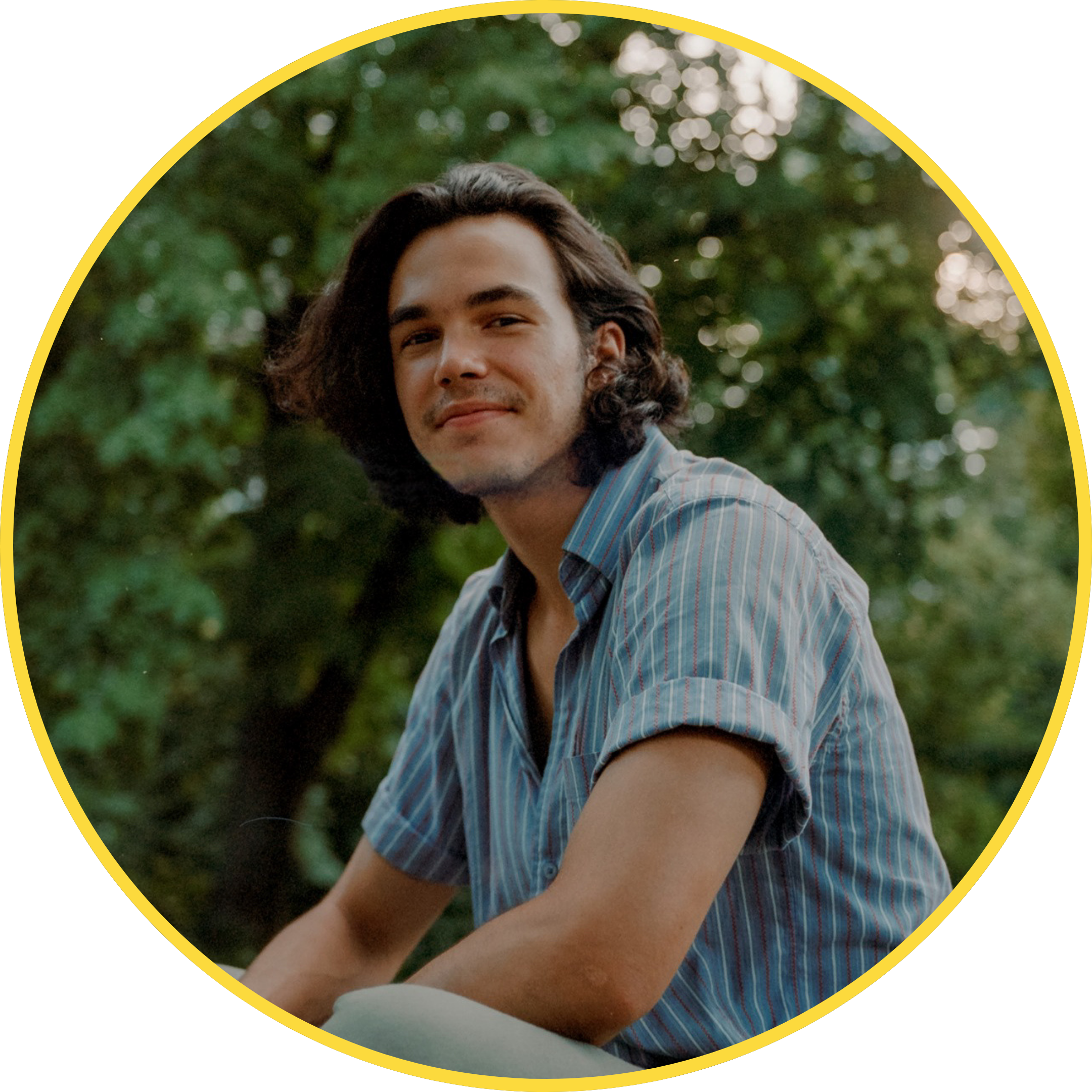 Headshot of a light-skinned twentysomething male of Latin American descent with dark brown, wavy, shoulder-length hair wearing a blue pin stripped shirt, sitting crossed-legged in front of luscious greenery.