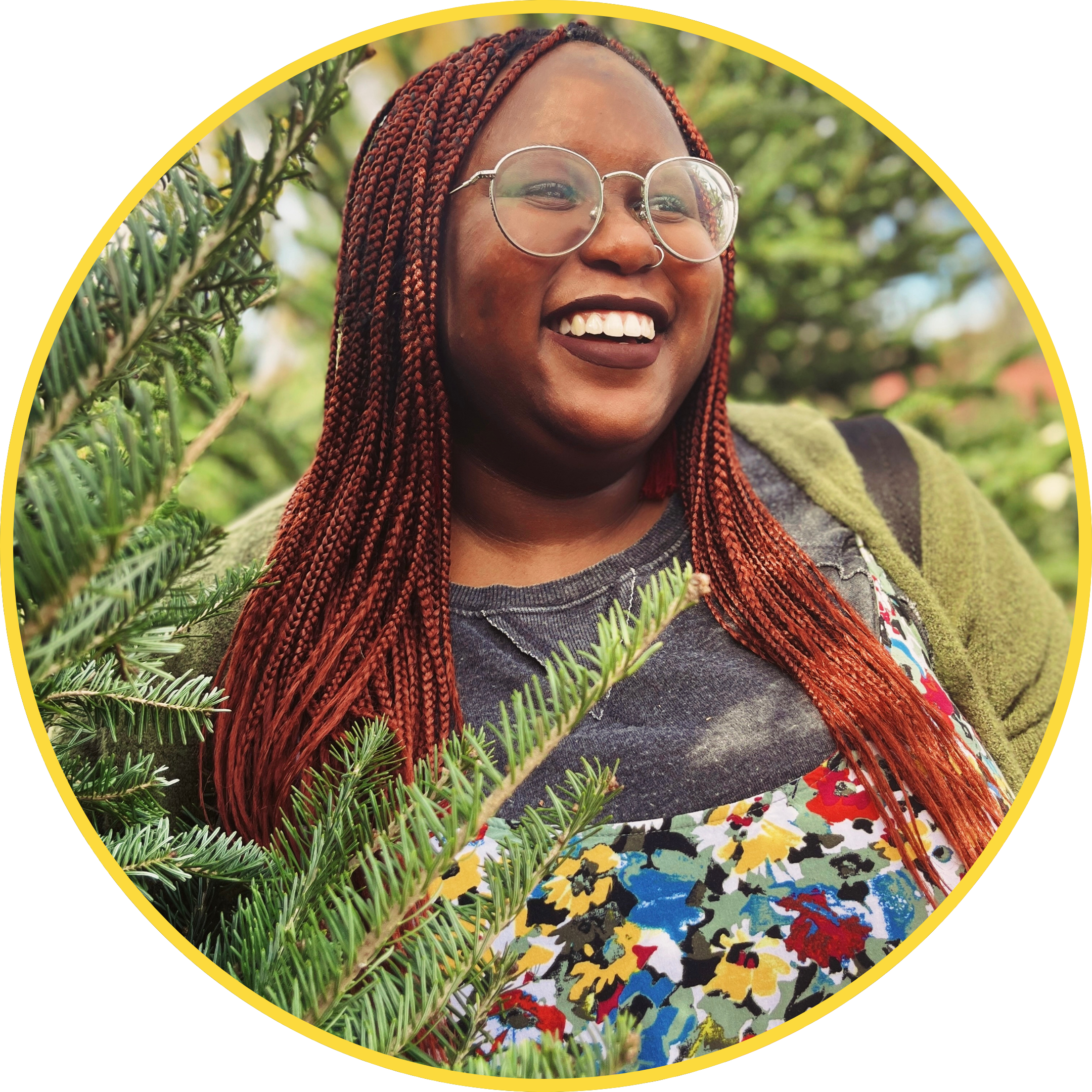 Headshot of a Black woman with round glasses, burnt orange box braids, wearing a floral jumpsuit and surrounded by green Christmas trees.