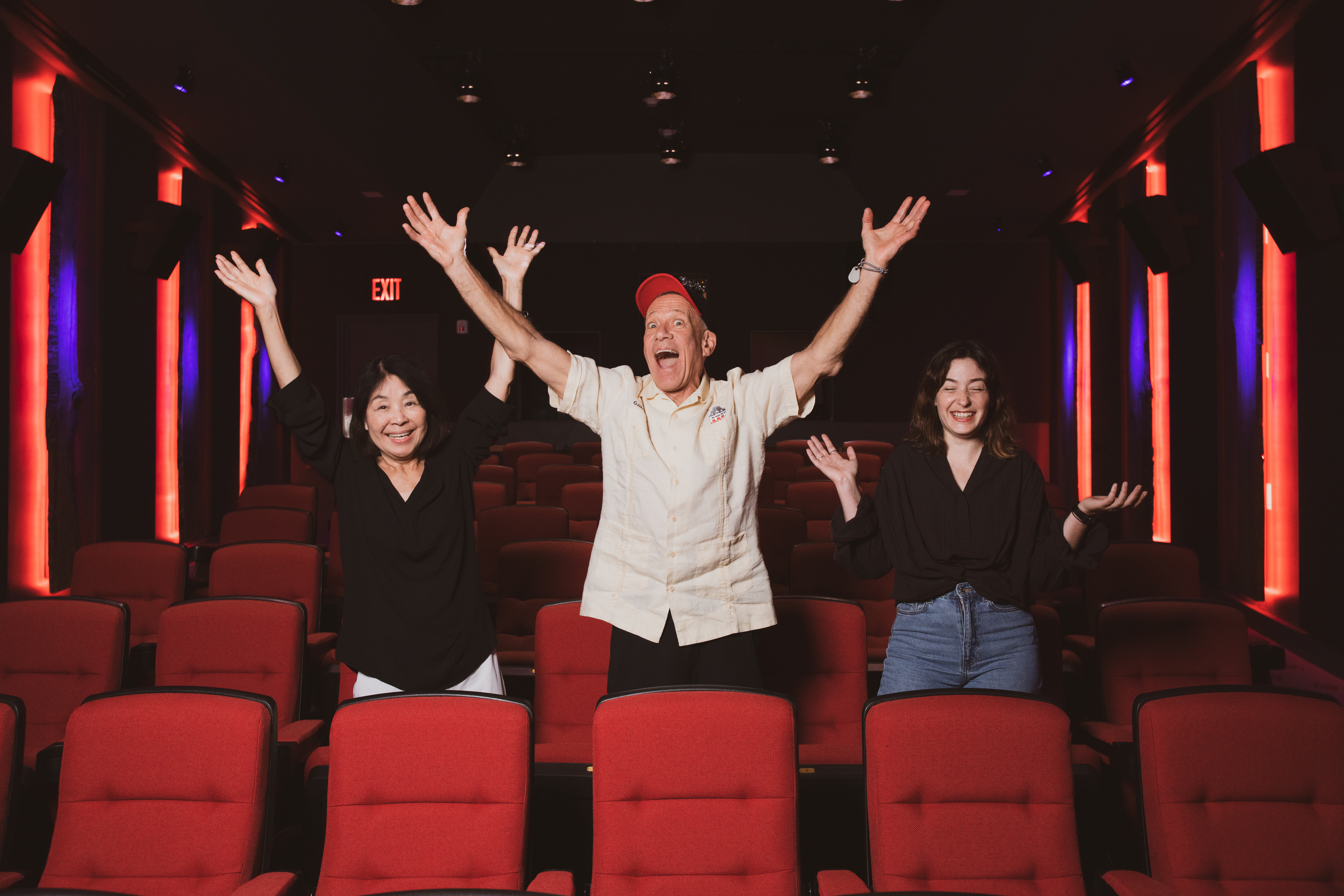 Three people stand with arms outstretched in a dramatically-lit cinema with red seats.