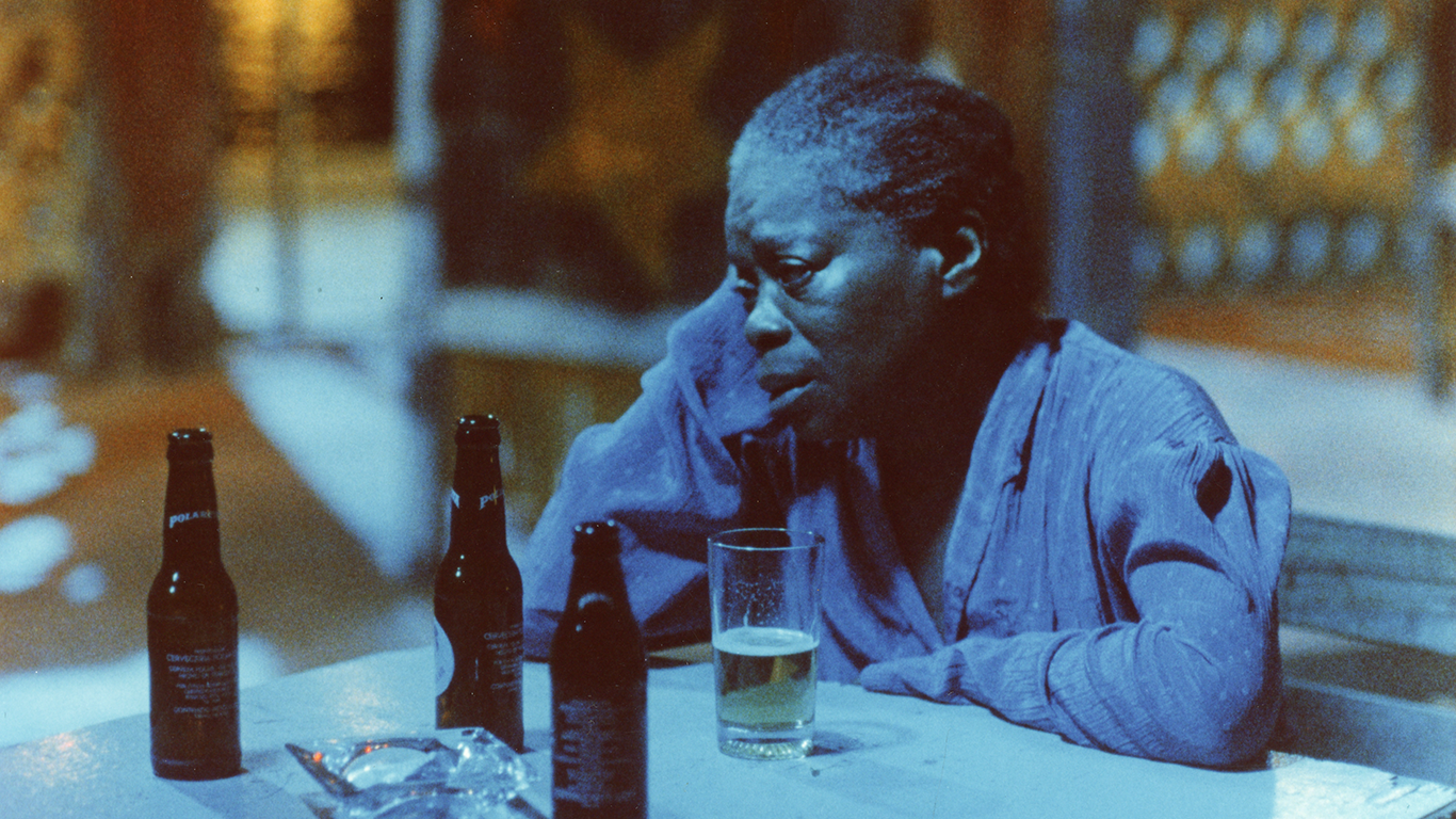 A woman leads her head on her right hand, while seated at a table drinking a beer.