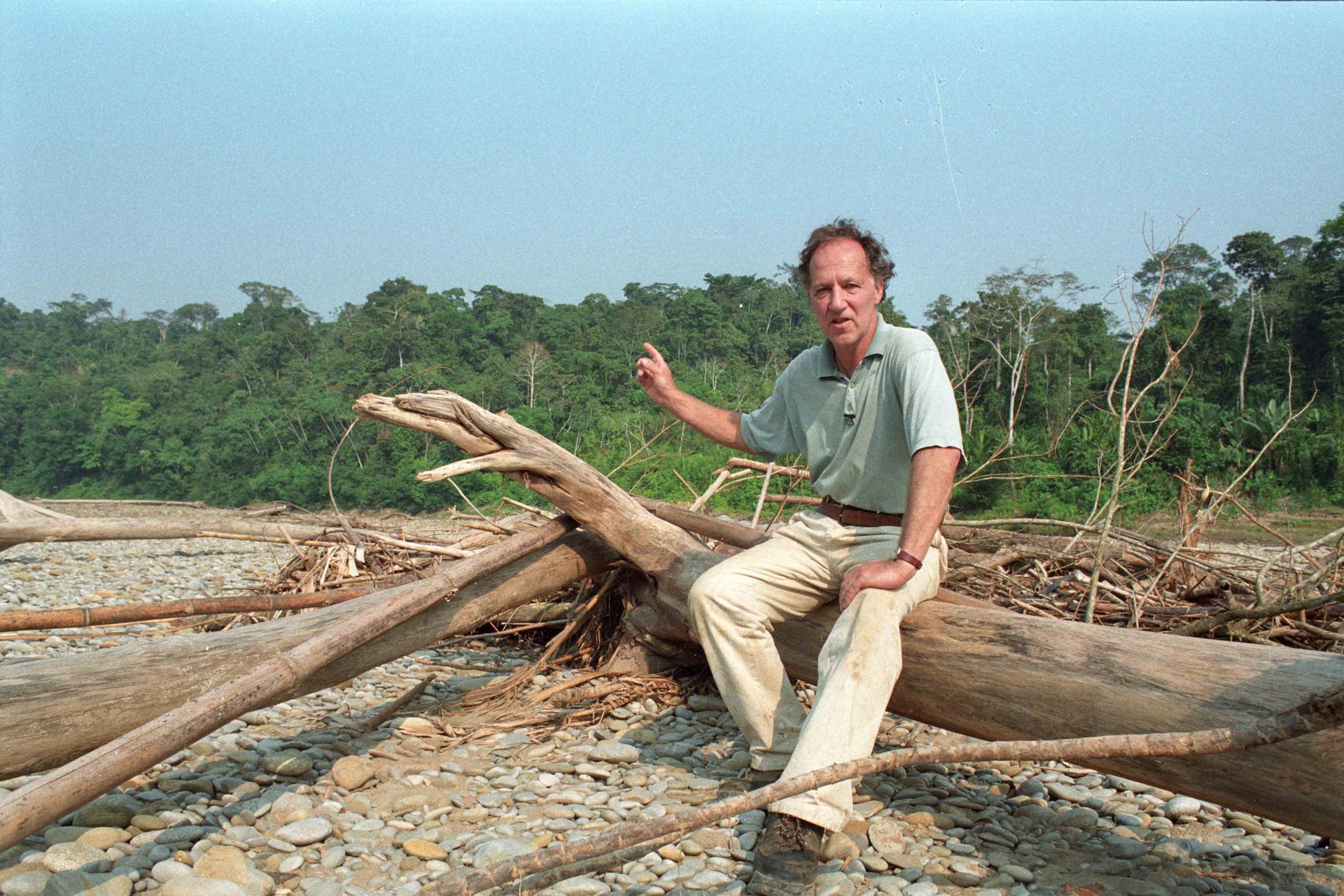 Werner Herzog sits on a large upturned, fallen tree.