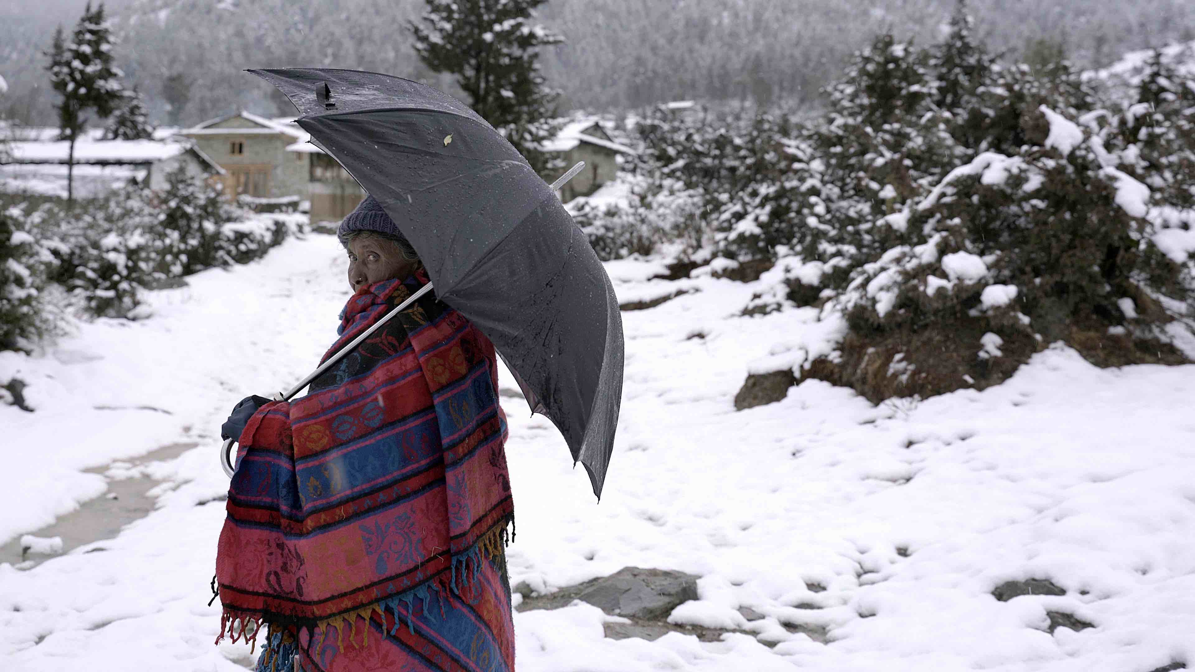 A woman with shawl wrapped around her, holding an umbrella, stands in a snowy environment. 