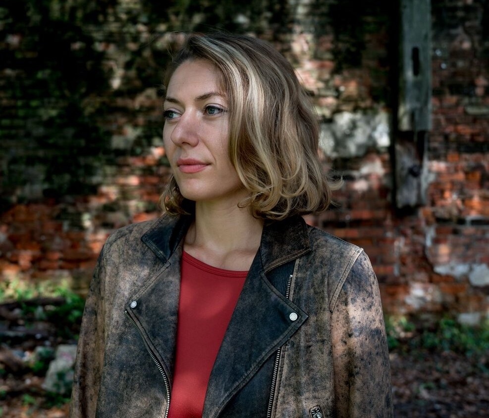 A white woman with blue eyes, brown hair and blonde streaks, looks off into the distance. She wears a red shirt and an acid-washed leather jacket. 