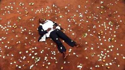 A black man in a white shirt, blue jacket, jeans, and barefoot lays on his back with his eyes closed. He is laying on a red dirt ground with white flowers and orange fruit scattered around him.