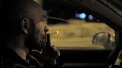 Profile of a white man with a shaved head sitting in a car at night, smoking