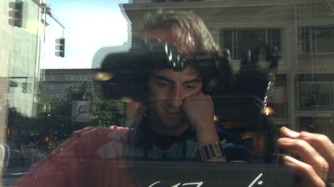 A young adult an through a window, looking at a laptop, with a reflection of a camera man