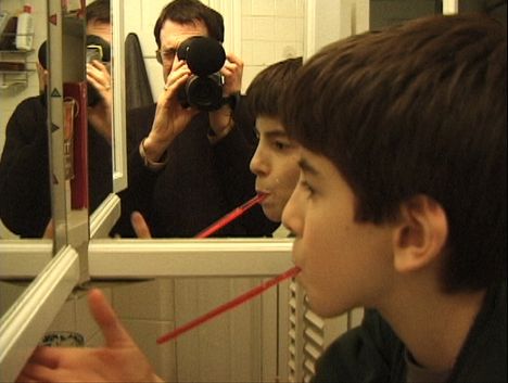 A young boy with dark hair brushes his teeth. In the mirror behind him is the reflection of an older man holding a video camera.