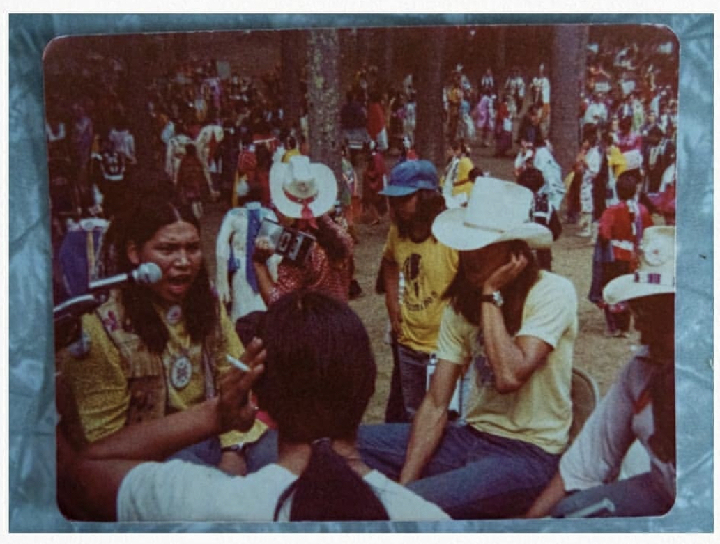 Film still from Powwow People. A group of Native American powwow participants sing in a circle as powwow attendees look on. 