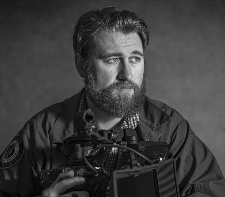 Black and white photo of person with short hair and beard, holding a camera. 