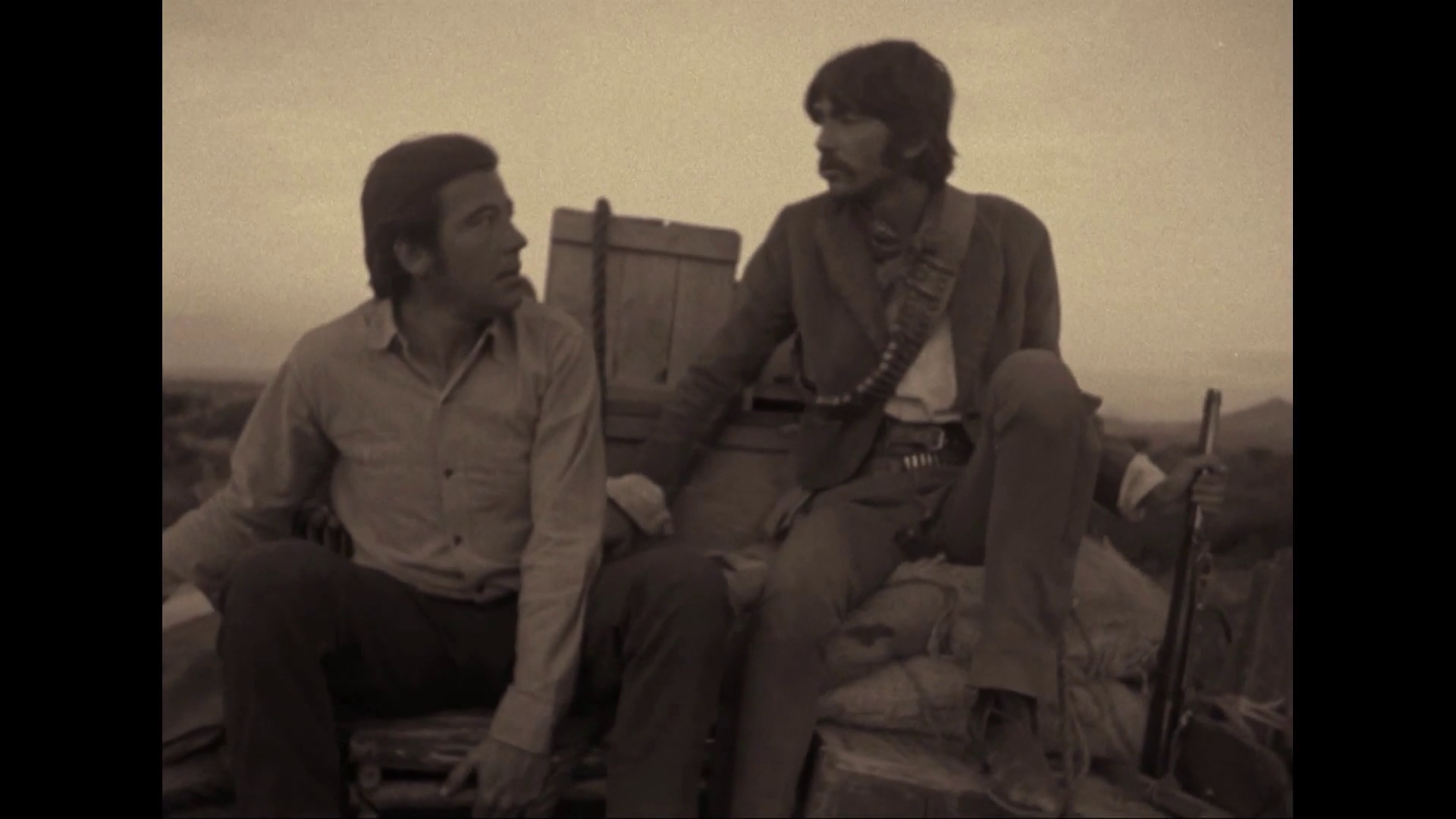 Two men sit on the back of a truck, looking at each other.