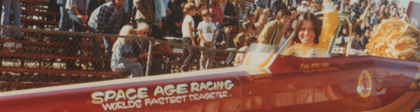 Archive photo of brown haired white woman Kitty O'Neil, race car driver, behind the wheel of red cigarette dragster car with painted words on it "space age racing worlds fastest dragster."