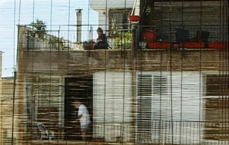 A man on his balcony and a woman on the balcony above, shot through blinds
