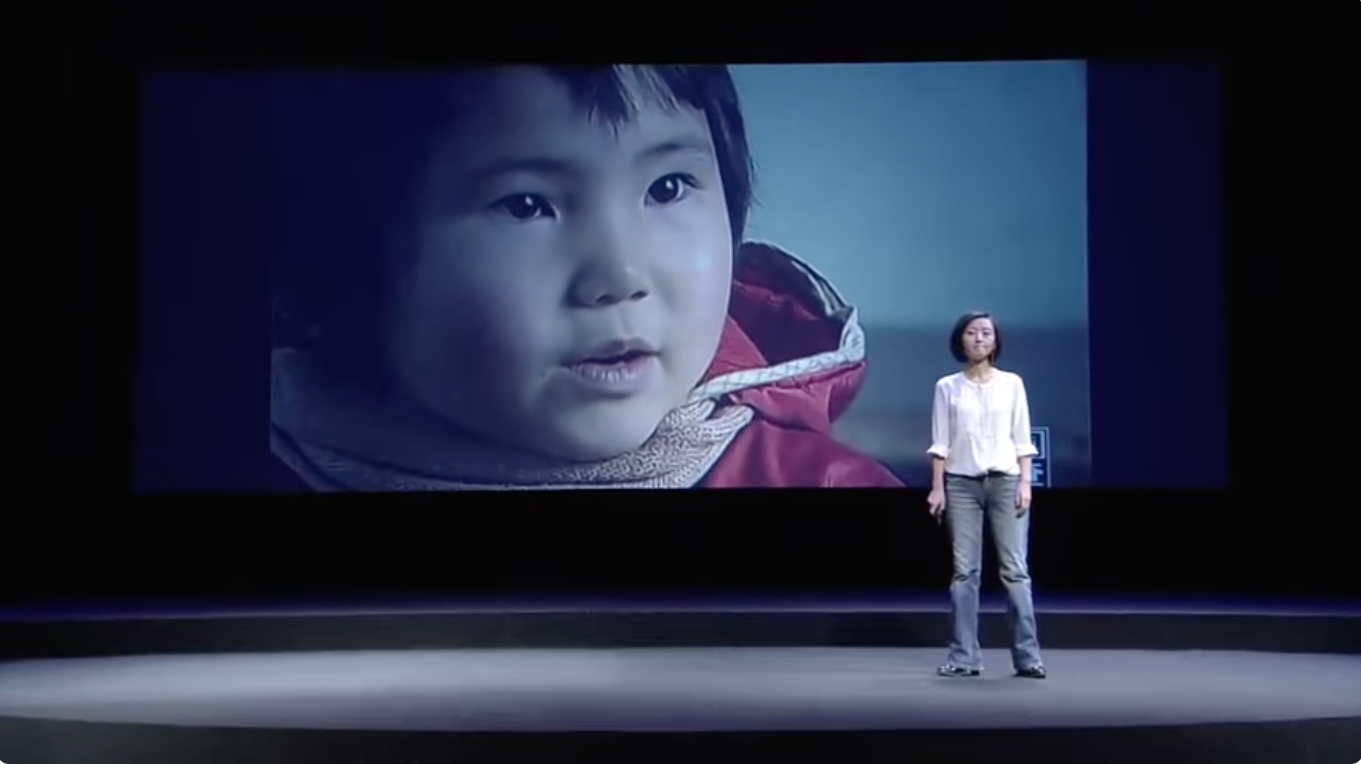 A journalist stands in front of a giant screen showing a young child.