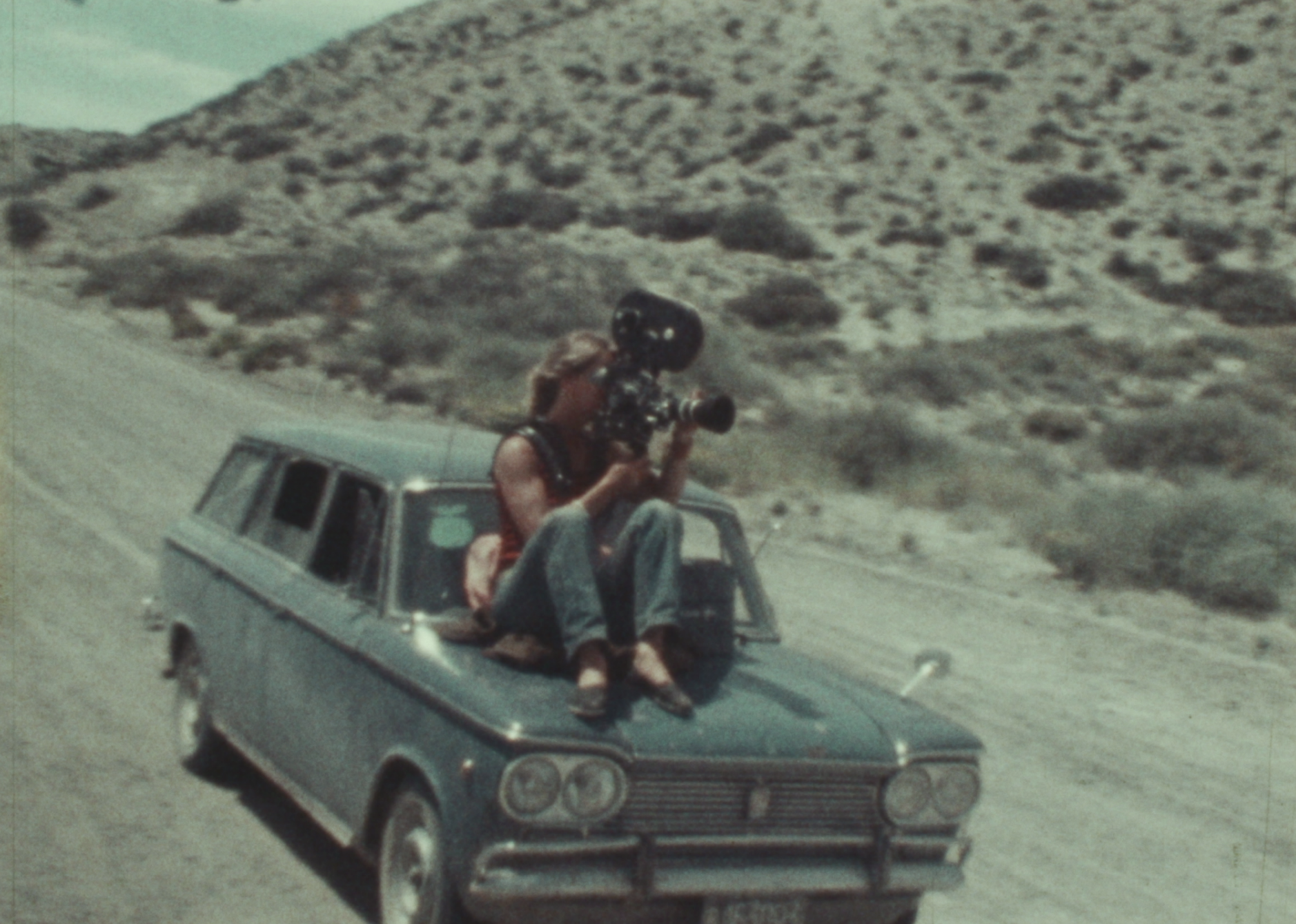 On top of a moving car, a woman sits holding a film camera.