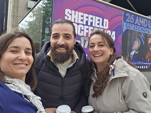 Two women stand on either side of a man, all smiling widely. The sign behind them is for Sheffield DocFest 2024.