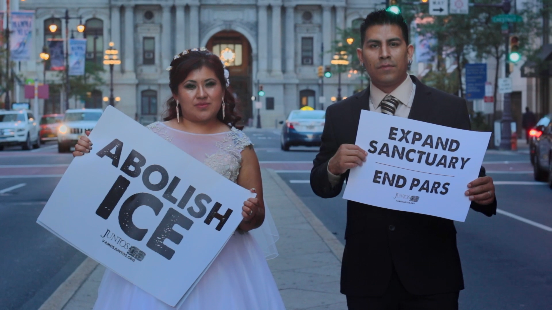 A film still from Expanding Sanctuary that shows a Latina woman wearing a white wedding gown and a Latino man wearing a black suit posing with blank expressions in front of Philadelphia City Hall holding posters that read "Abolish ICE" and "Expand Sanctuary, End PARS".