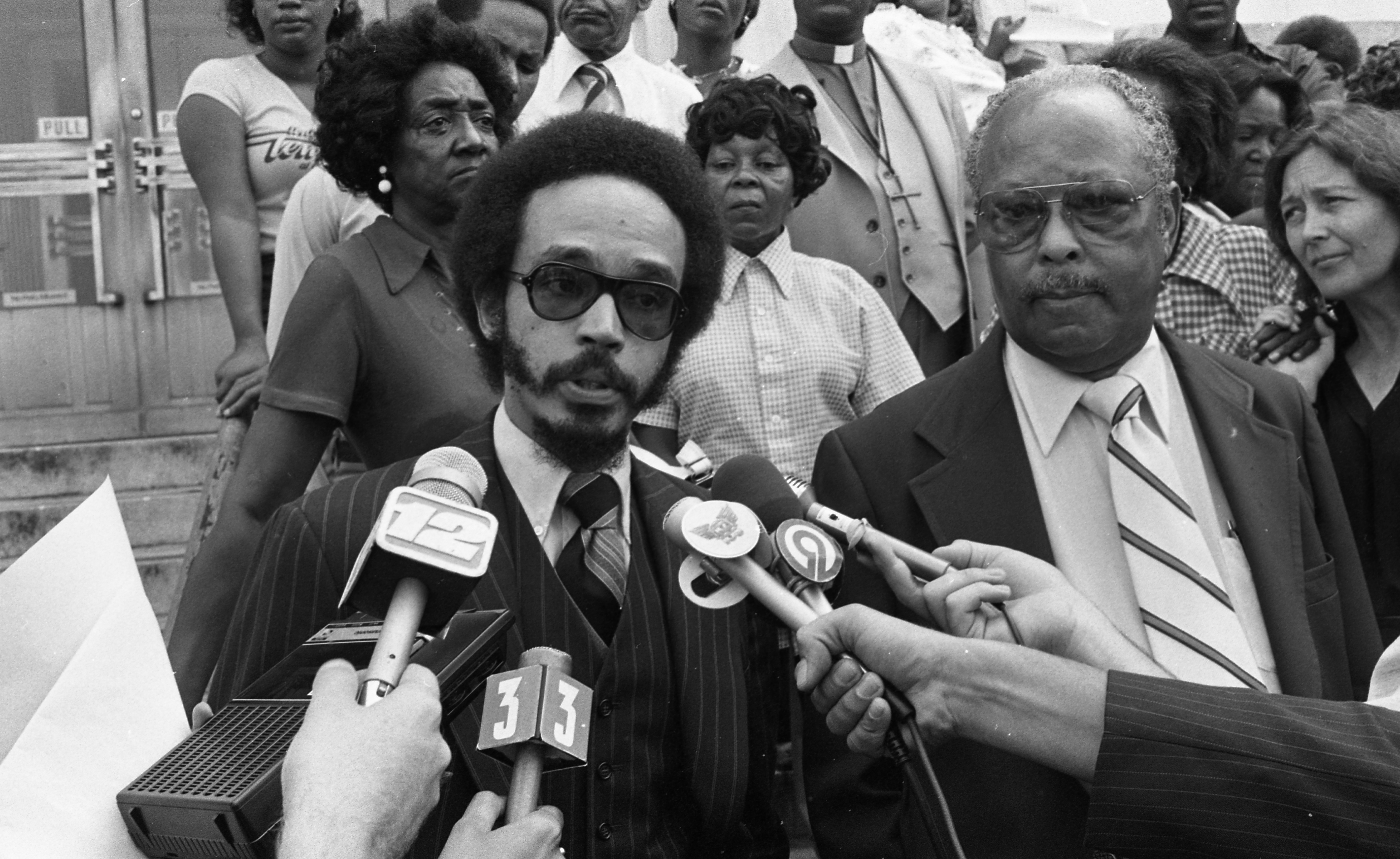 A film still from How to Sue the Klan that shows a black-and-white photo of a young Black man speaking at a press conference with several reporters microphones pointed towards him. Directly next to the man is another Black man with a serious expression, and behind them are a predominantly Black audience all with blank and serious facial expressions.