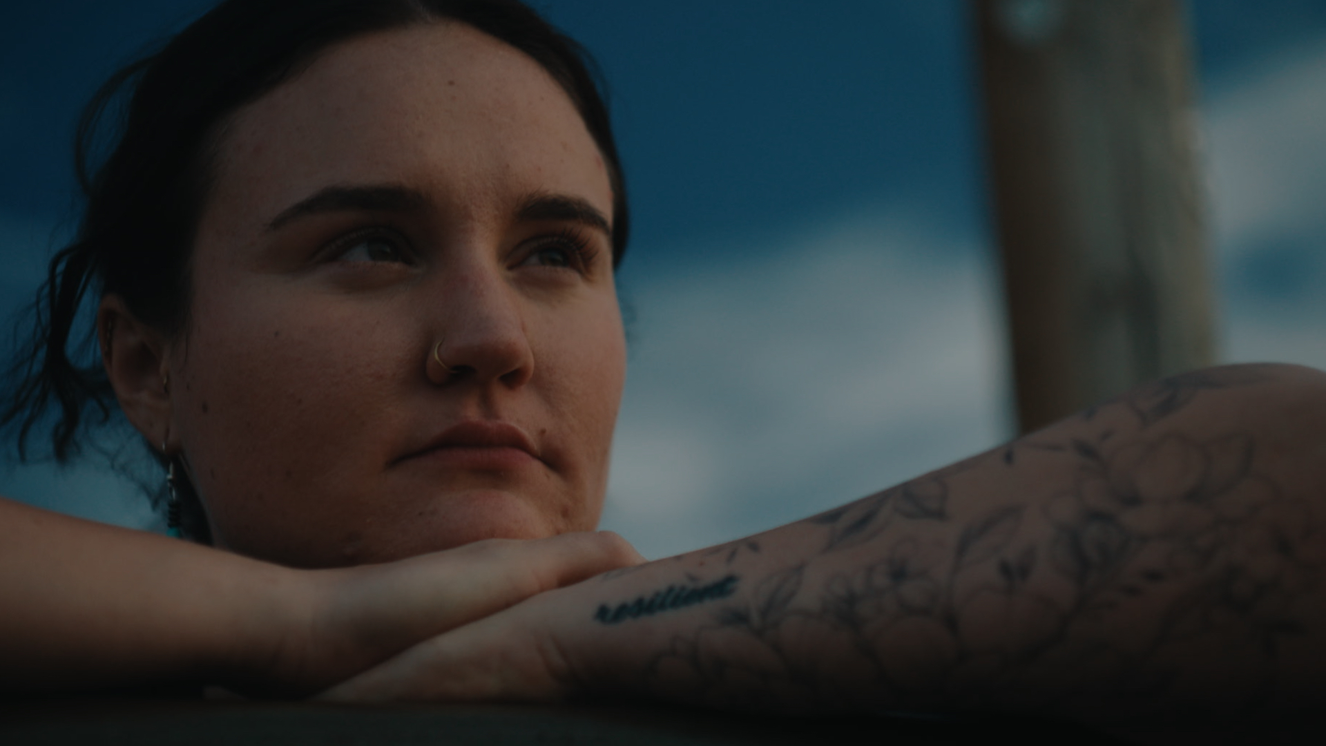 A film still from Of Medicine and Miracles that shows a young woman with a nose ring looking thoughtfully off to the side. Her arms, one adorned with dainty floral tattoos, are crossed atop each other, with her head gently resting on her hands against a dark blue sky.