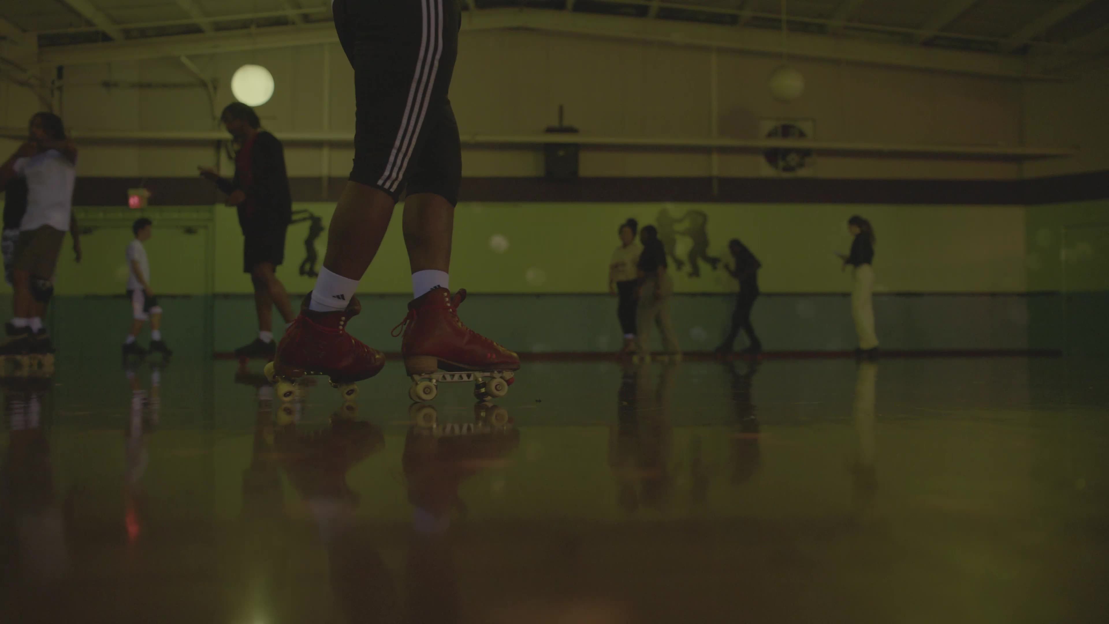 A film still from The People Could Fly that shows a person in an indoor roller skating rink wearing white socks, adidas black striped joggers and red roller skaters from the waist down. As they glide across the floor there are several other skaters visible in the dimly lit background.