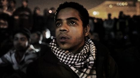 A young man wearing a black and white scarf stands in a crowd at night, looking in the distance