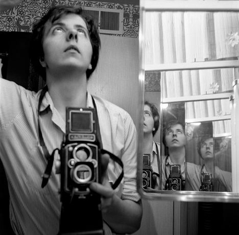 A black and white image of a woman with short hair and a camera held up to her chest, taken in a mirror with many repeating images in the reflection