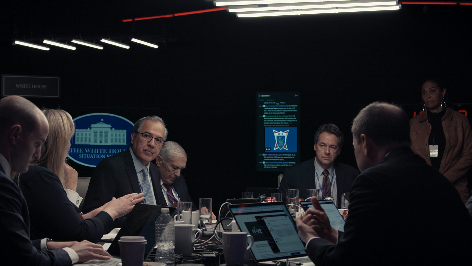 A group of people are meeting over compiter screens in front of them. A sign behind them reads ‘The White House’.