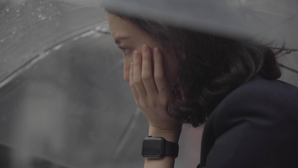 Film still of a pensive Shiori under an umbrella, face in hand.