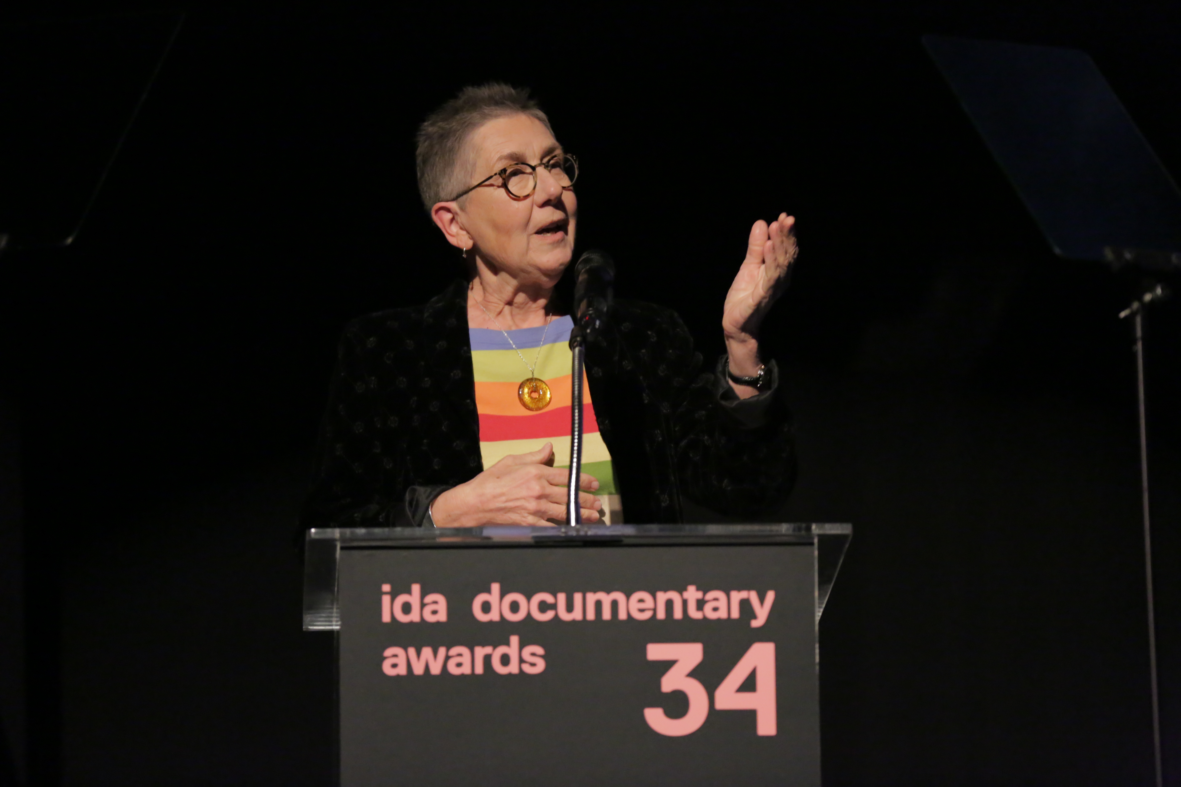 Julia Reichert, a white woman with grey hair and glasses, wearing a striped shirt beneath a dark blazer, at the podium accepting the 2018 IDA Career Achievement Award. Photo: Rebecca Sapp