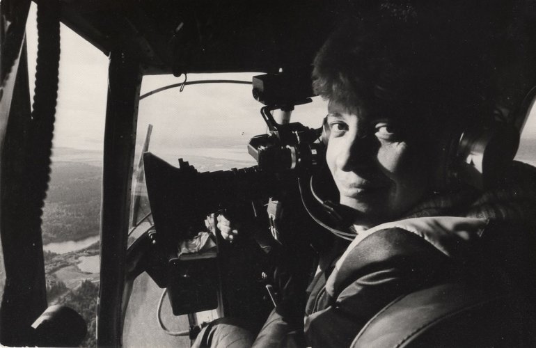 Black and white image of a woman in a helicopter looking back at the camera as she holds a camera