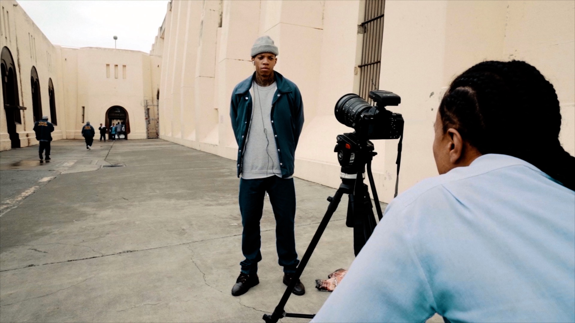 Images courtesy of "What These Walls Won't Hold" film: A photograph of a young Black man standing in front of a camera on a tripod being interviewed, with the filmmaker's back in the foreground.
