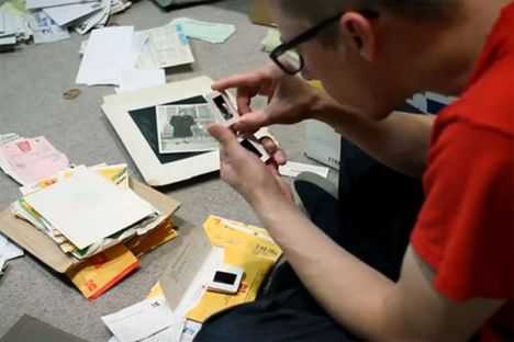 A man in a red tshirt and glasses sits on the floor looking at small proofs, with piles of pictures and documents around him