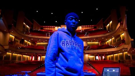 A young black man in a Harvard University sweatshirt stands with his DJ setup and an ornate empty theater behind him