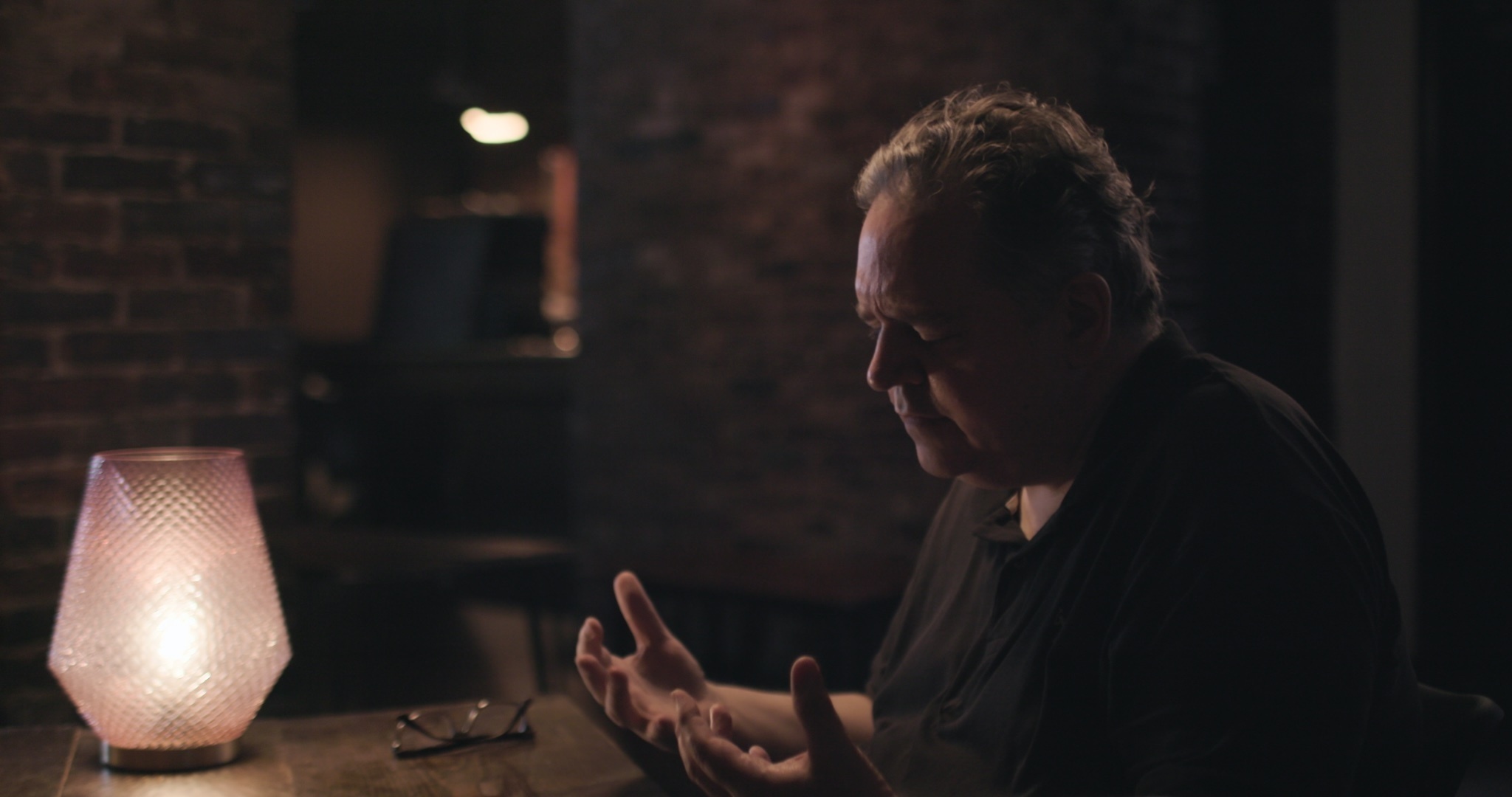 A man sits in a dark room, lit by a lamp next to him, staring at his hands.