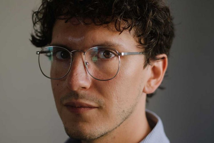 A portrait shot of Gerardo del Valle in front of a gray wall. He is wearing a dress shirt, wears glasses, and has short curly brown hair)