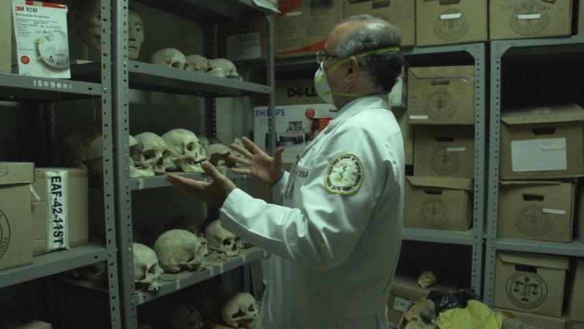 Still from 'The Room of Bones,' depicting a man in a white lab coat staring at shelves full of human remains.
