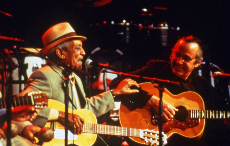 An elder Cuban musician playing guitar and singing on stage next to another man with a guitar.