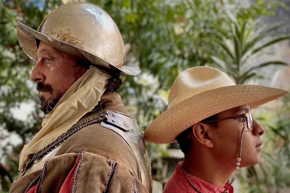 Actor Eduardo San Juan Breña is a Mexican man with a beard, dressed as the Conquistador. He is standing next to musician Tomás de Aquino, who is wearing a red shirt, a straw hat and glasses, with a beaded suspender. Image from Rodrigo Reyes’ “499." Courtesy of the filmmaker.
