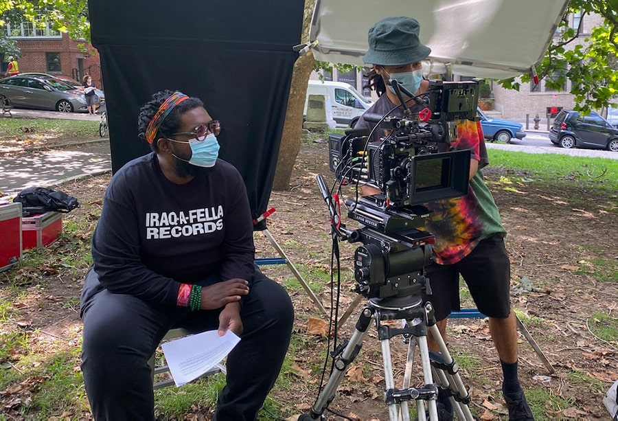 Filmmaker Ahmir "Questlove" Thompson, a Black man in a colorful headband and black tshirt sitting, and DP Shawn Peters, a Black cameraperson with a green hat, on the sets of “Summer of Soul.” Courtesy of Searchlight Pictures.