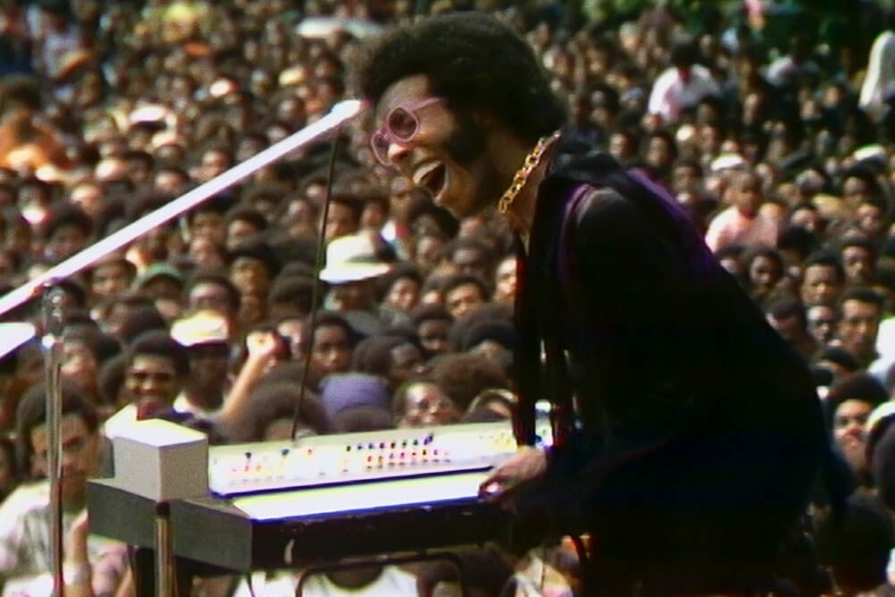 Sly Stone, a Black male musician in a velvet coat singing to a large audience at the Harlem Cultural Festival. Courtesy of Searchlight Pictures.