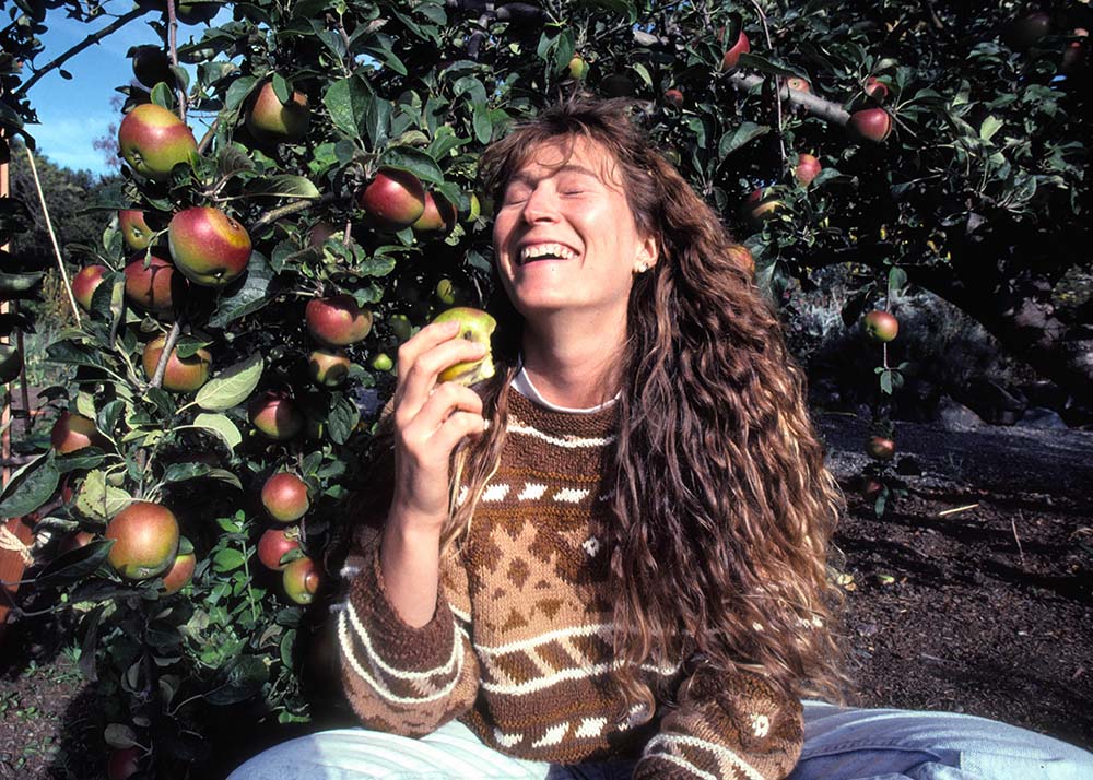 Image of a smiling white woman in a brown sweater with long hair, sitting under an apple tree. From Mark Kitchell’s 2017 film 'Evolution of Organic.' Courtesy of Mark Kitchell.