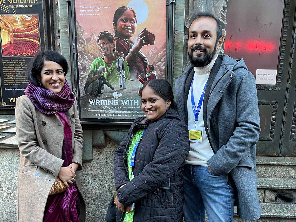 Protagonist Meera Devi standing in front of the poster for 'Writing With Fire' with filmmakers Rintu Thomas and Sushmit Ghosh, at IDFA 2021. Courtesy of Rintu Thomas and Sushmit Ghosh.