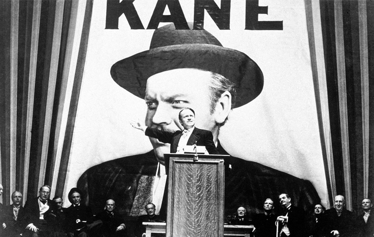 A man stands on a podium with a giant poster that reads 'Kane' in the back. Most first-time viewers of 'Citizen Kane'  don't realize that the News on the March short that opens the film is a clever parody of a series of theatrical shorts produced by Time, Inc. 