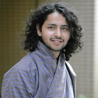 Head shot of a male of south Asian descent with brown skin and long curly black hair. He is wearing a blue striped 'Gho' which is the national dress of Bhutan.