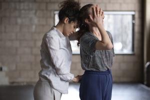 This photograph shows dancer Mor Mendel closely behind dancer Hadar Ahuvia, the two facing towards the right in an industrial looking dance studio with windows. They hold opposite hands behind Hadar's back, while the other pair of opposite hand's covers Hadar's eyes. Hadar's head leans back into Mor's forehead— they are contemplative. 