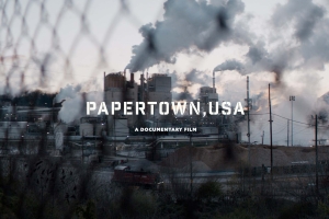 A factory seen from behind a wire fence, the text "Papertown, USA - A Documentary Film" is superimposed on the image in white 